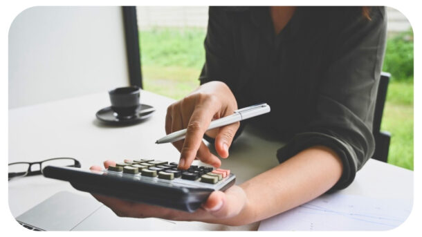 A person using a calculator while holding a pen, with a coffee cup, glasses, and documents on a desk.
